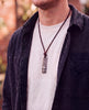 Man in a dark overshirt and white tee wearing a cross bar necklace on a brown leather cord, rustic Christian necklace for men