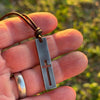 Cutout cross bar pendant for men held in the hand outdoors to show size, rustic gunmetal stainless steel on leather cord