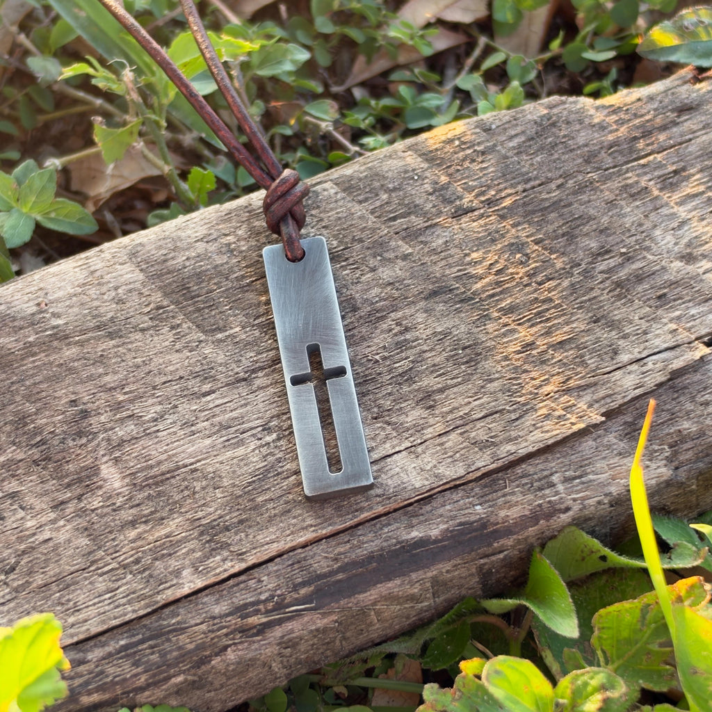 Mens cutout cross bar pendant on a leather cord hanging over weathered wood among green leaves, rustic Christian jewelry
