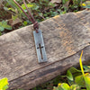 Mens cutout cross bar pendant on a leather cord hanging over weathered wood among green leaves, rustic Christian jewelry