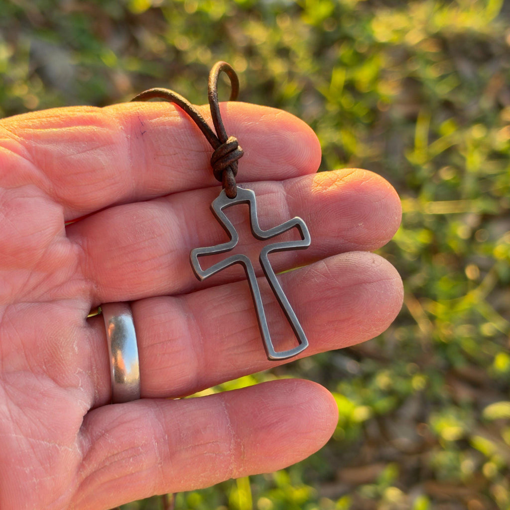 Open cross necklace for men held in a man's hand outdoors to show its size, handmade stainless steel surfer cross on leather cord