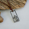 Surfer cross necklace for men, The Breakthrough Cross in dark stainless steel with the cross breaking out of the frame, on a brown leather cord resting on beach driftwood