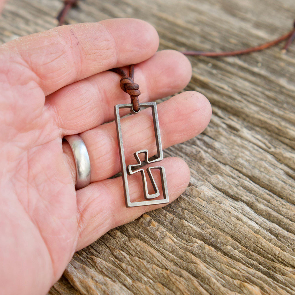 Men's cross necklace held in the hand to show size, handmade stainless steel surfer cross pendant on a leather cord