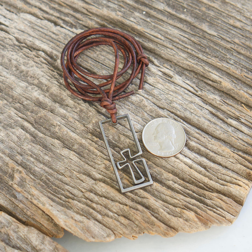 Stainless steel cross pendant for men beside a coin for scale, surfer cross necklace with coiled brown leather cord on weathered wood