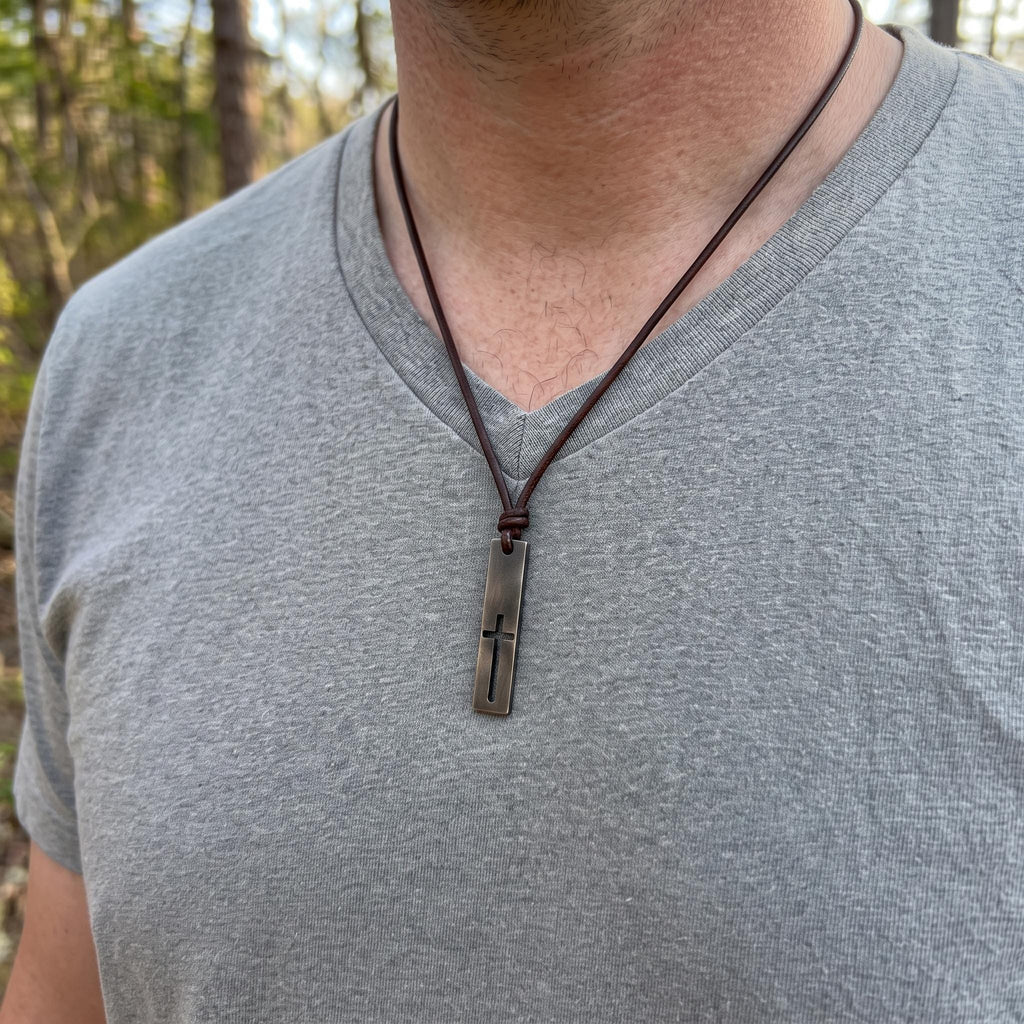 Man in a gray t-shirt wearing the cutout cross bar necklace on a brown leather cord, a rustic cross necklace for men