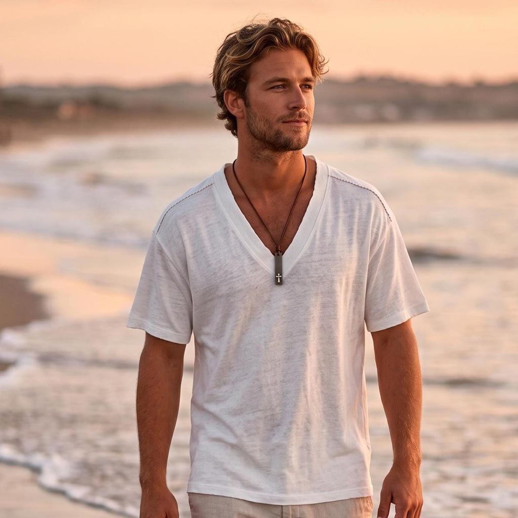 Man in a white t-shirt on the beach at sunset wearing a stainless steel cross bar necklace on a leather cord, surfer cross for men