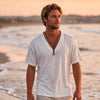 Man in a white t-shirt on the beach at sunset wearing a stainless steel cross bar necklace on a leather cord, surfer cross for men