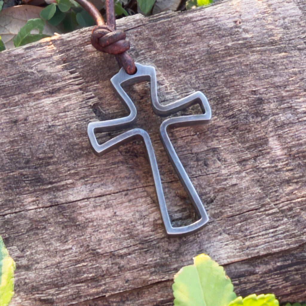 Stainless steel open cross pendant for men on a brown leather cord resting on weathered driftwood, rustic beach jewelry