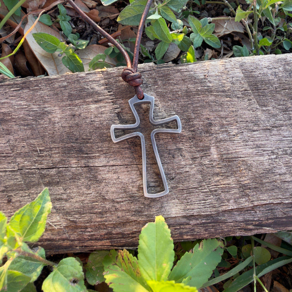 Surfer cross necklace for men hanging over a weathered wooden log among green leaves, open stainless steel cross on leather cord